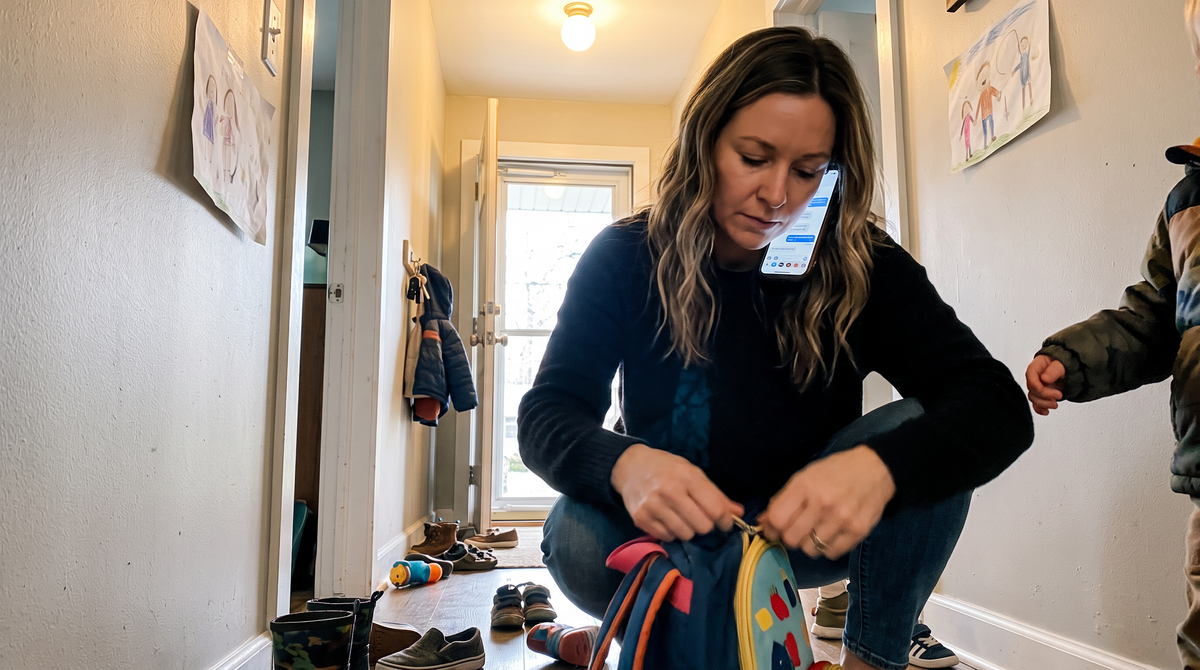 A woman crouched in her front hallway zipping a child's backpack, phone pinned to her shoulder